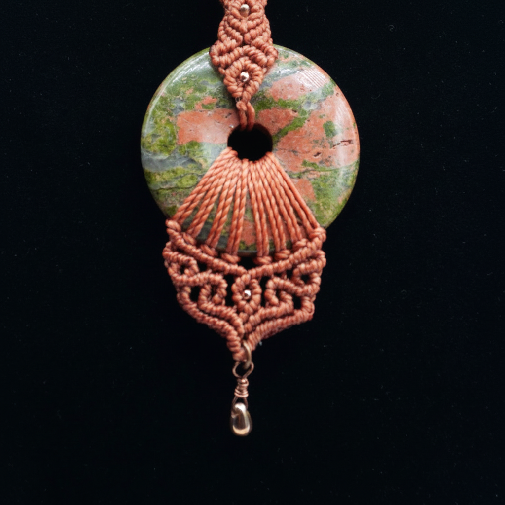 A handmade macrame pendant featuring a donut shape Unikite stone disc with a cutout, adorned with intricate terracotta red braided cord and a small clear bead dangle, set against a solid black background.