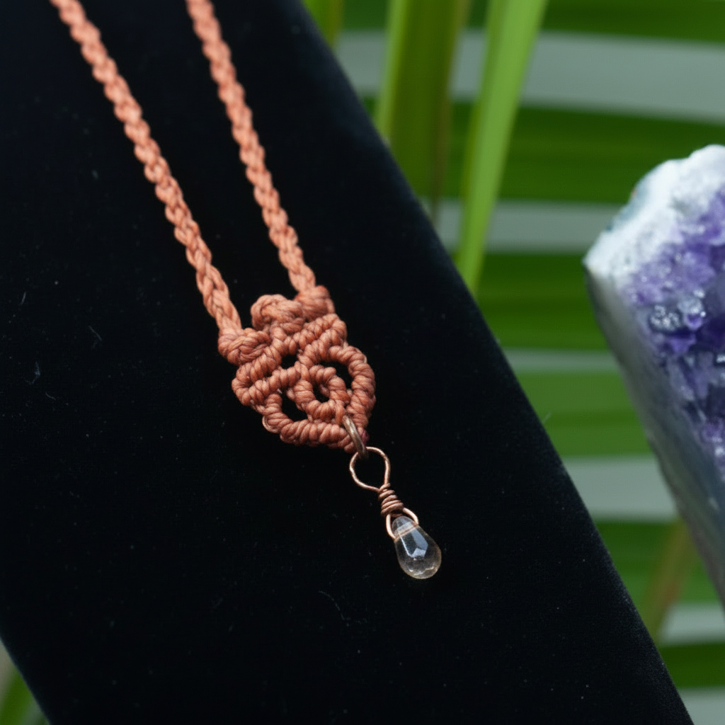 A close-up of a handmade macrame necklace in a terracotta red color, featuring a decorative knot and a small clear glass bead with a copper wire wrap, displayed on a black velvet jewelry stand.