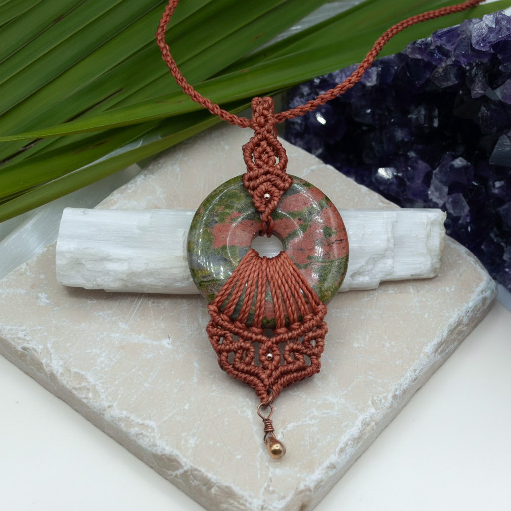 A handmade pendant necklace with a terracotta red macrame cord and a donut Unikite stone pendant, styled on a stone slab with a white selenite rod and purple amethyst cluster.