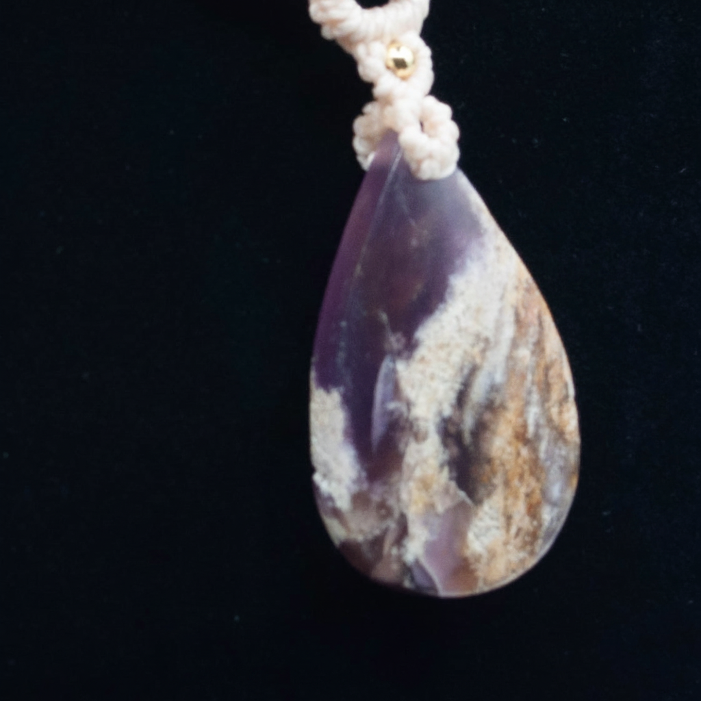 A teardrop-shaped Amethyst gemstone pendant with a marbled pattern of purple, cream, and brown, suspended from a ivory macrame knotted cord against a black background.