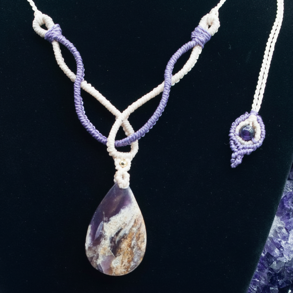 A handmade macrame necklace with a teardrop-shaped amethyst pendant, displayed on a black velvet jewelry bust. The necklace features interwoven ivory and purple cords, with a matching purple macrame charm on the necklace end shown on the right.