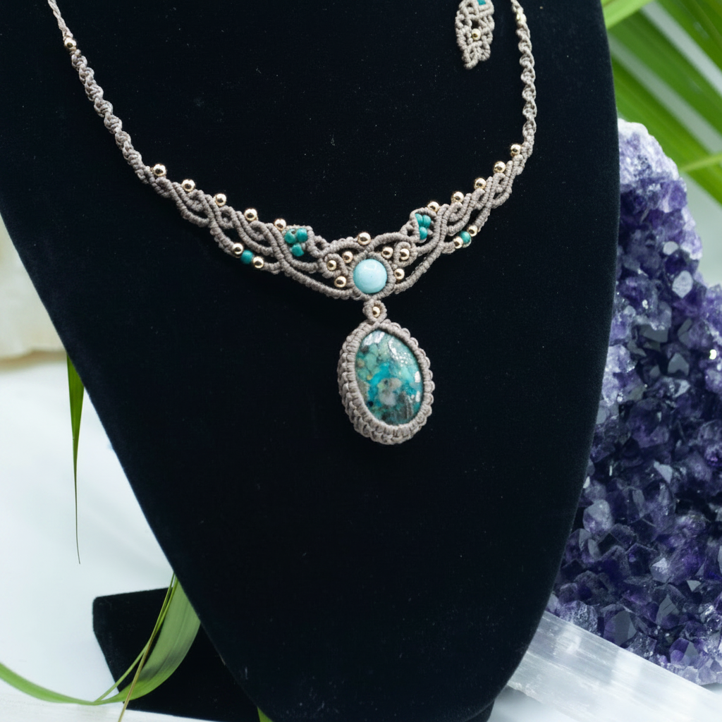 A taupe brown macrame necklace with turquoise and blue-green stones, displayed on a black jewelry bust. A matching small pendant or charm is visible at the top right, and decorative green leaves and a purple crystal cluster frame the display.