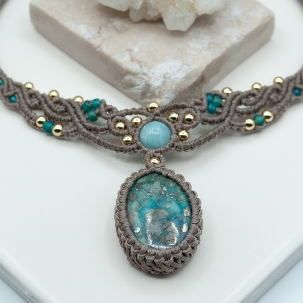 A close-up of a handmade macrame necklace with a large oval pendant. The necklace is woven in taupe-brown cord with small gold beads and turquoise-colored accent beads.