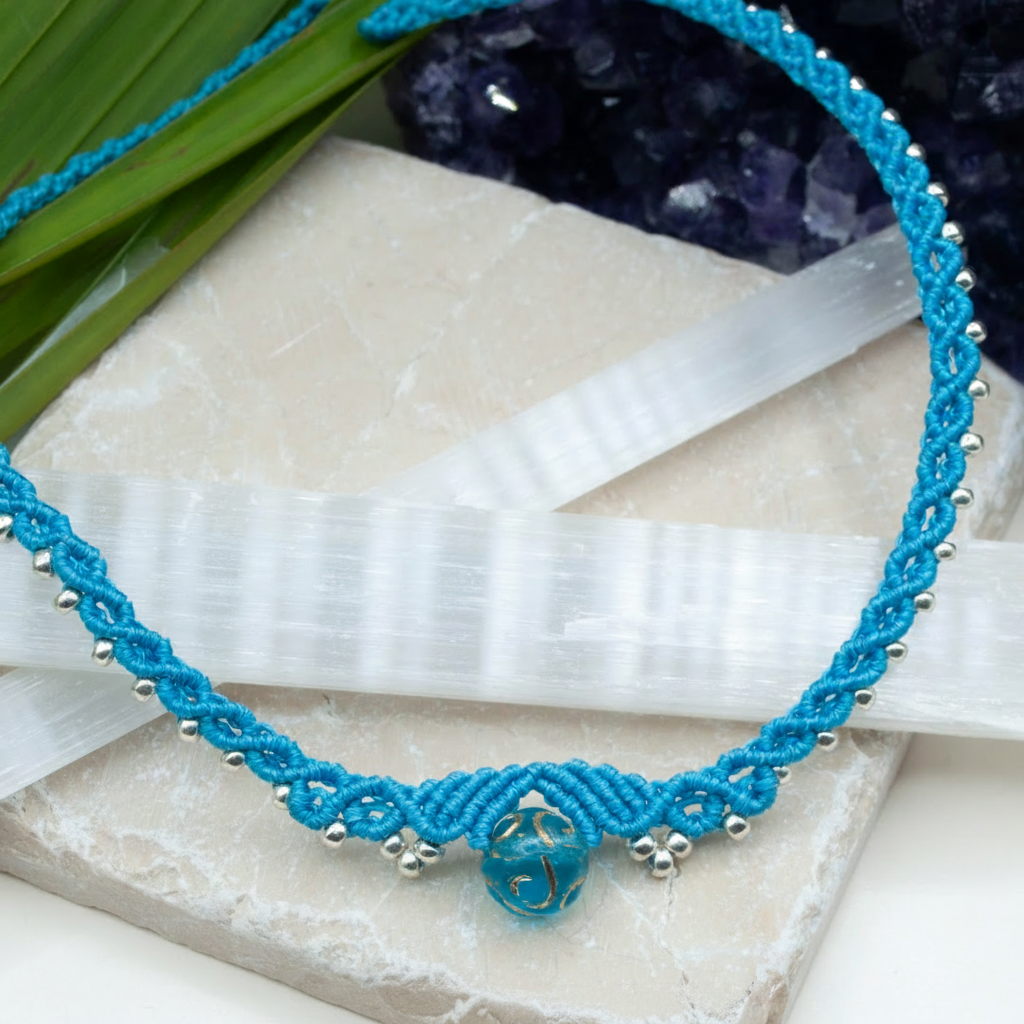 A turquoise blue macrame necklace with a central blue glass bead and silver-toned metal accents, displayed on white crystal slices with green leaves and purple crystals in the background.