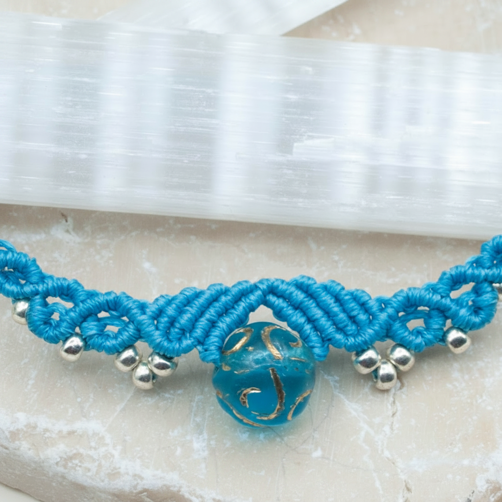 A turquoise blue macrame necklace with a central round blue glass bead featuring gold swirls and small silver metal beads, displayed on a light stone surface with a white crystal rod in the background.