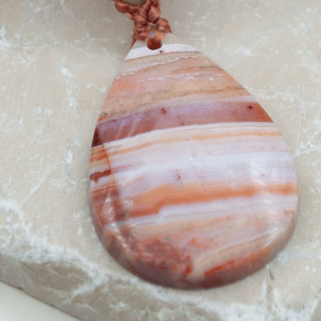 A teardrop-shaped pendant made of polished striped agate or jasper stone in shades of pink, orange, cream, and brown, hanging from a rust cord. The pendant is displayed on a textured beige stone surface.