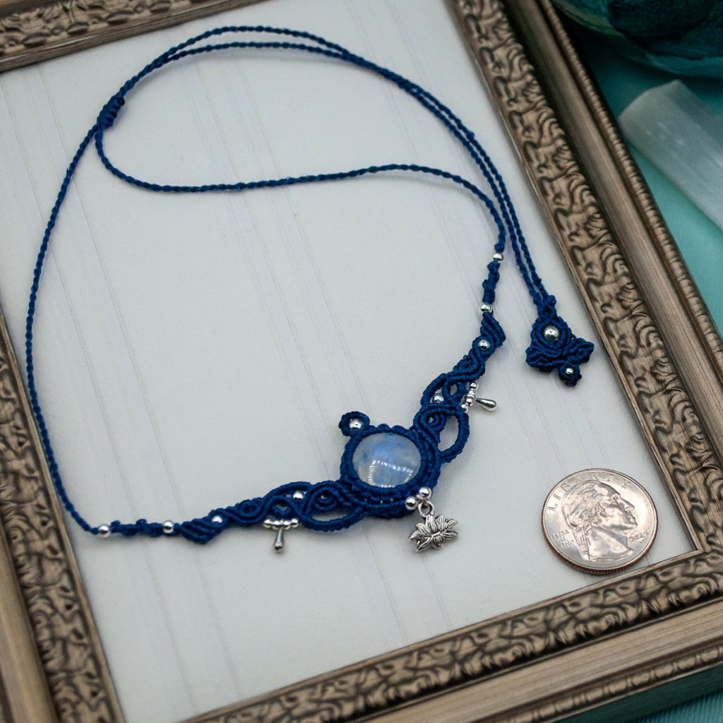 A handmade macrame necklace in dark blue cord with a central round moonstone and small silver charms, displayed on a white surface inside an ornate gold frame next to a coin for scale.
