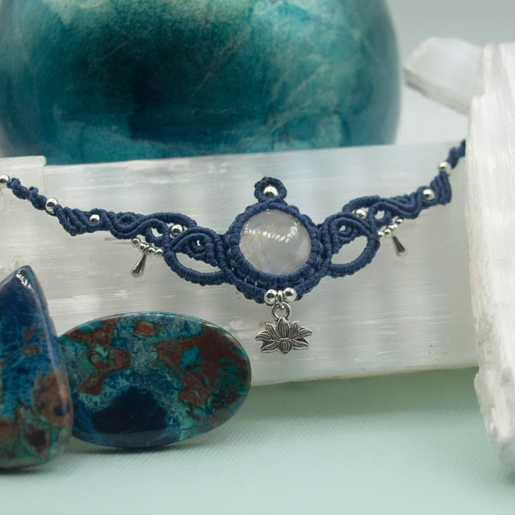 A handmade macrame necklace with a central white stone and a silver lotus charm, displayed on a white selenite slab with decorative stones in the background.