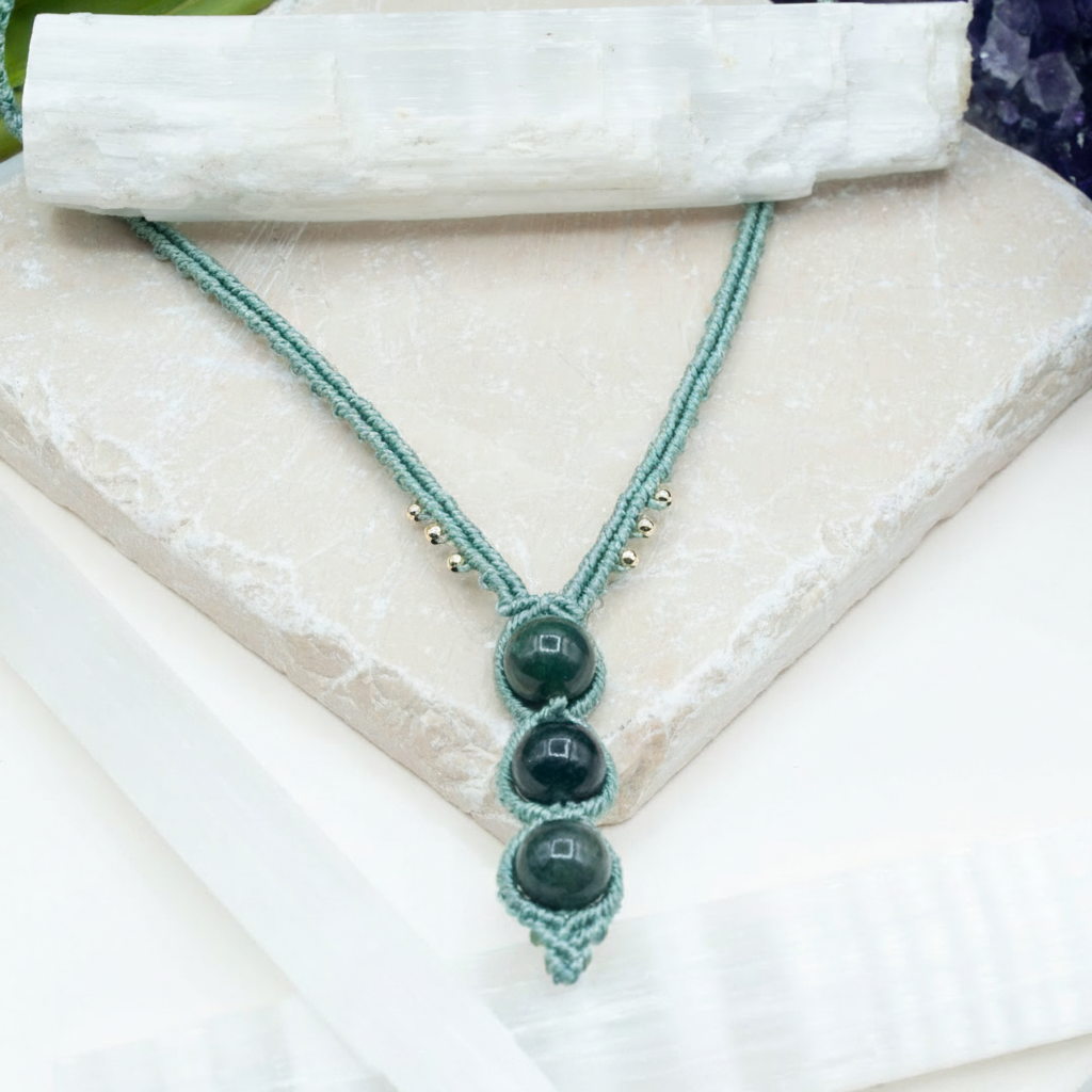 A handmade macrame necklace with three dark green polished beads, displayed on a white stone slab with a white selenite wand prop. The necklace has a mint green cord and small gold spacer beads.