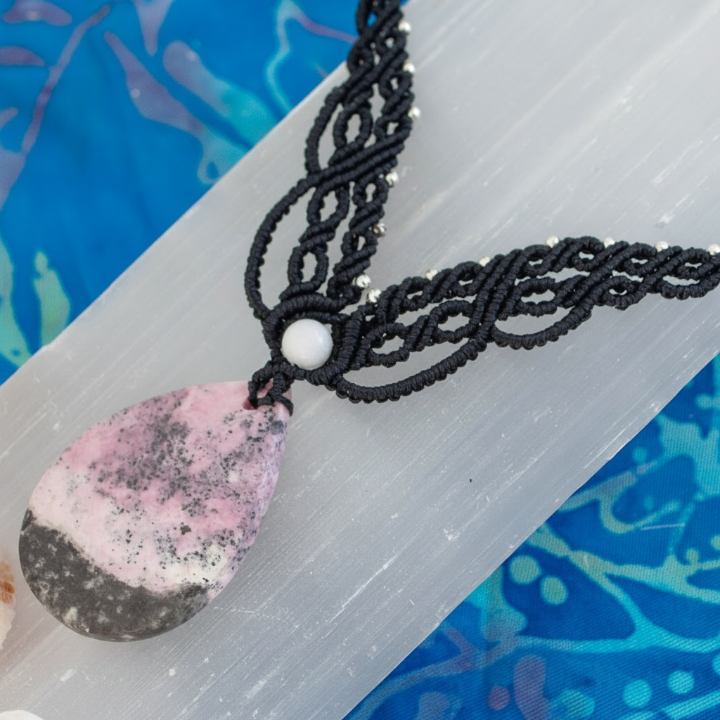 A close-up of a handmade macrame necklace with a pink and black stone pendant and a white bead, displayed on a translucent white selenite rod over a blue patterned background.