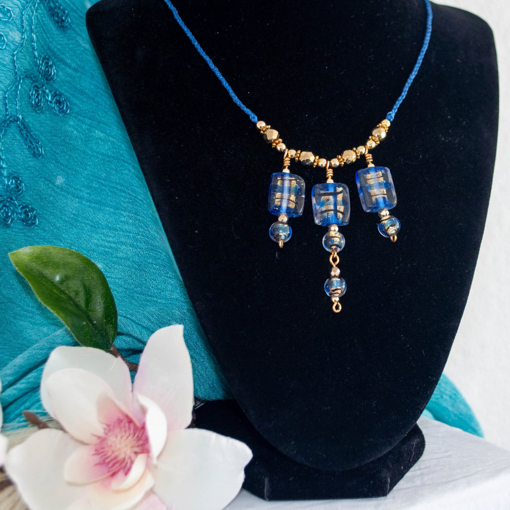 Handmade cobalt blue lampwork glass macrame necklace with gold foil detail, three rectangular focal beads, gold-tone bead accents, and four-strand braided cord, displayed on black velvet jewelry bust with teal fabric and magnolia flower.
