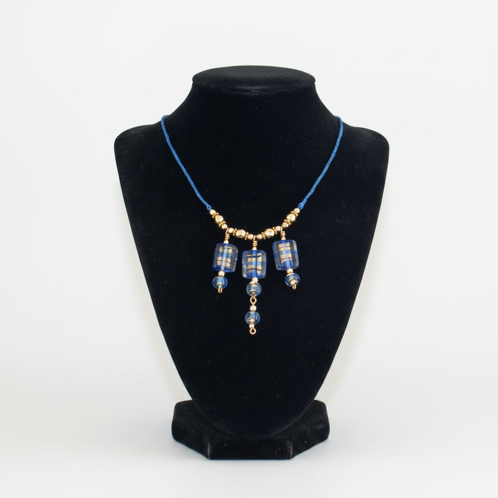 Handmade cobalt blue lampwork glass macrame necklace with gold foil detail, three rectangular focal beads, gold-tone and hematite bead accents, and four-strand braided cord, displayed on black velvet jewelry bust against grey background.