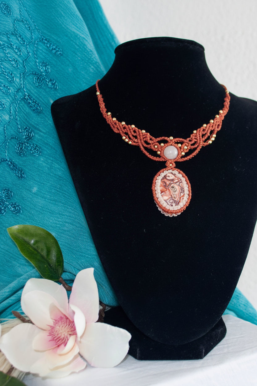 A handmade terracotta red braided cord necklace with a carved oval pendant, displayed on a black velvet jewelry bust. The necklace features intricate macramedetails and a central white pearl accent.