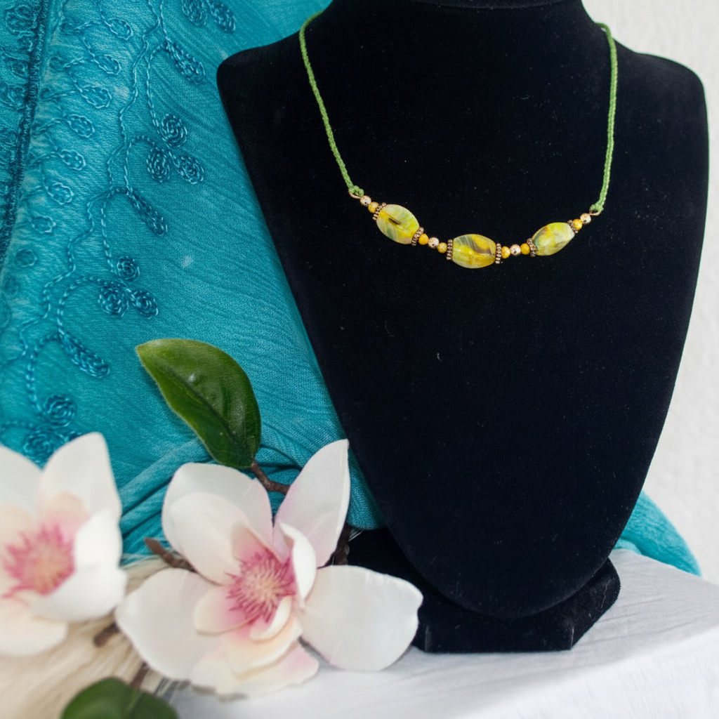 Handcrafted green cord macrame necklace with twisted rectangle chartreuse lampwork glass beads on a brass wire bar and small brass spacer beads, displayed on a black velvet jewelry bust with white flowers and teal embroidered fabric backdrop.