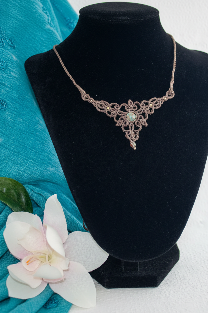 A handmade macrame necklace in a mocha brown color, featuring a central round calsilica stone, displayed on a black velvet jewelry bust. The background includes a bright teal fabric drape and a white surface.