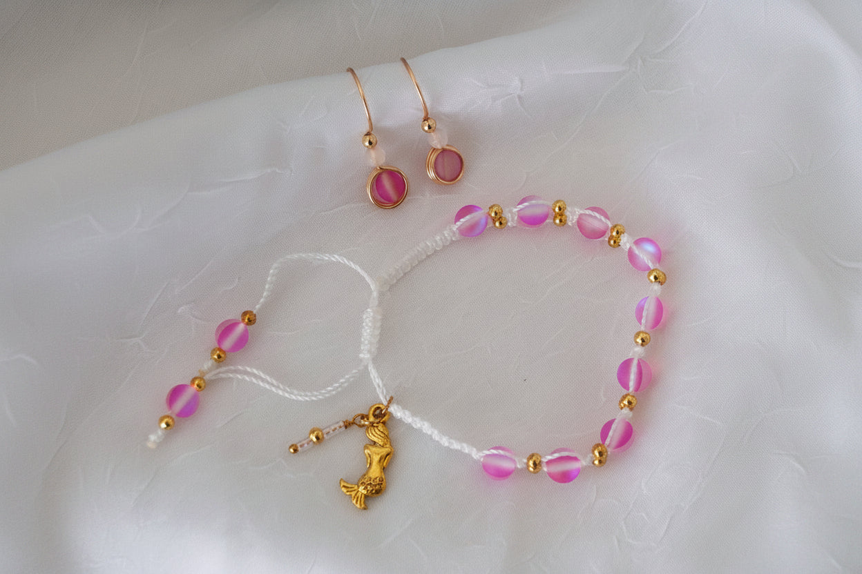 A handmade pink mermaid glass macrame bracelet with brass spacer beads and a gold brass mermaid charm, styled on white linen alongside matching pink cat's eye drop earrings with rose gold hooks. Each item sold separately.