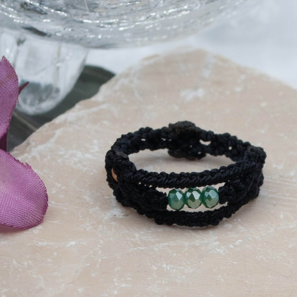 A handmade macrame band ring in black flat waxed nylon cord with a double-wrap braided design and three faceted mint green glass crystal beads across the center face, displayed on a rose quartz surface with a soft-focus pink flower and glass vessel in the background.