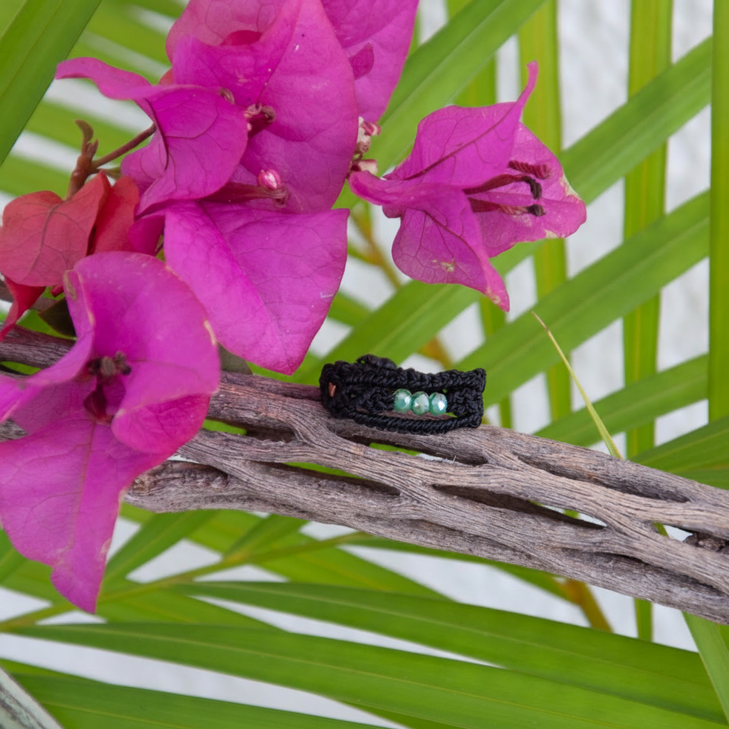 A handmade macrame band ring in black flat waxed nylon cord with three faceted mint green glass crystal beads, resting on a piece of driftwood surrounded by vibrant magenta bougainvillea flowers and tropical palm fronds