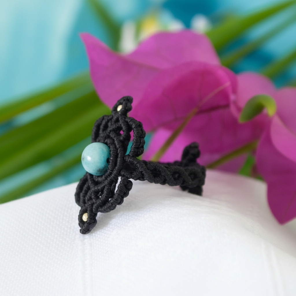 A side angle view of a handmade bohemian macrame ring in black flat waxed nylon cord with an intricate knotwork design and round Amazonite bead center, displayed on a white surface with a magenta orchid and tropical teal background.