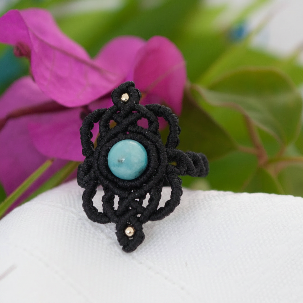 A handmade bohemian macrame ring in black flat waxed nylon cord with an intricate lace-like knotwork design, featuring a round Amazonite bead center and small brass accent beads, displayed on a white surface with a soft-focus magenta flower background.