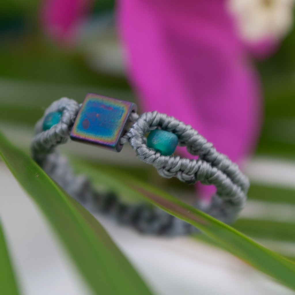 A side angle view of a handmade minimalist gray macrame ring with an iridescent square Miyuki Tila bead at the center and vibrant teal seed beads, resting on a tropical green leaf with a magenta flower in the soft-focus background.