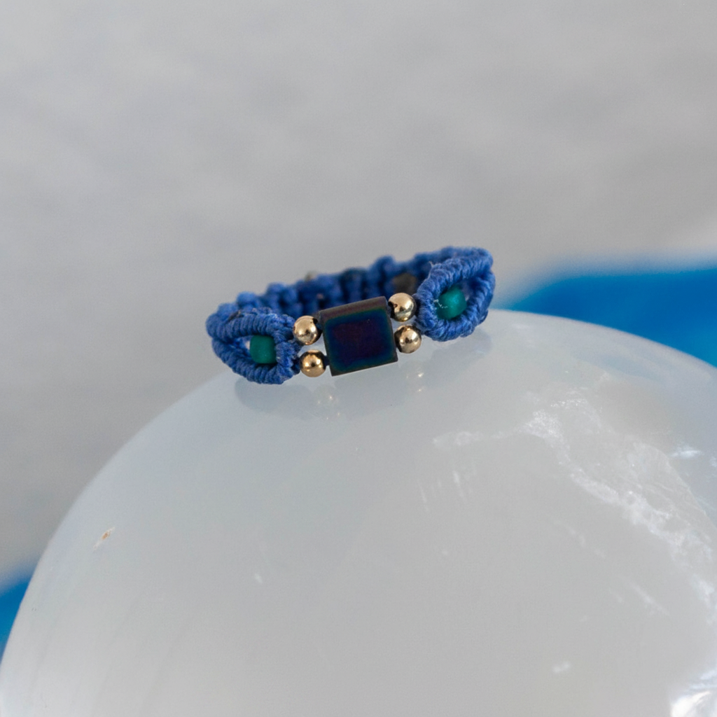 A handmade minimalist periwinkle macrame ring with a square Miyuki Tila bead center, teal seed beads, and gold brass accent beads, balanced on top of a white selenite sphere with a soft blue background.