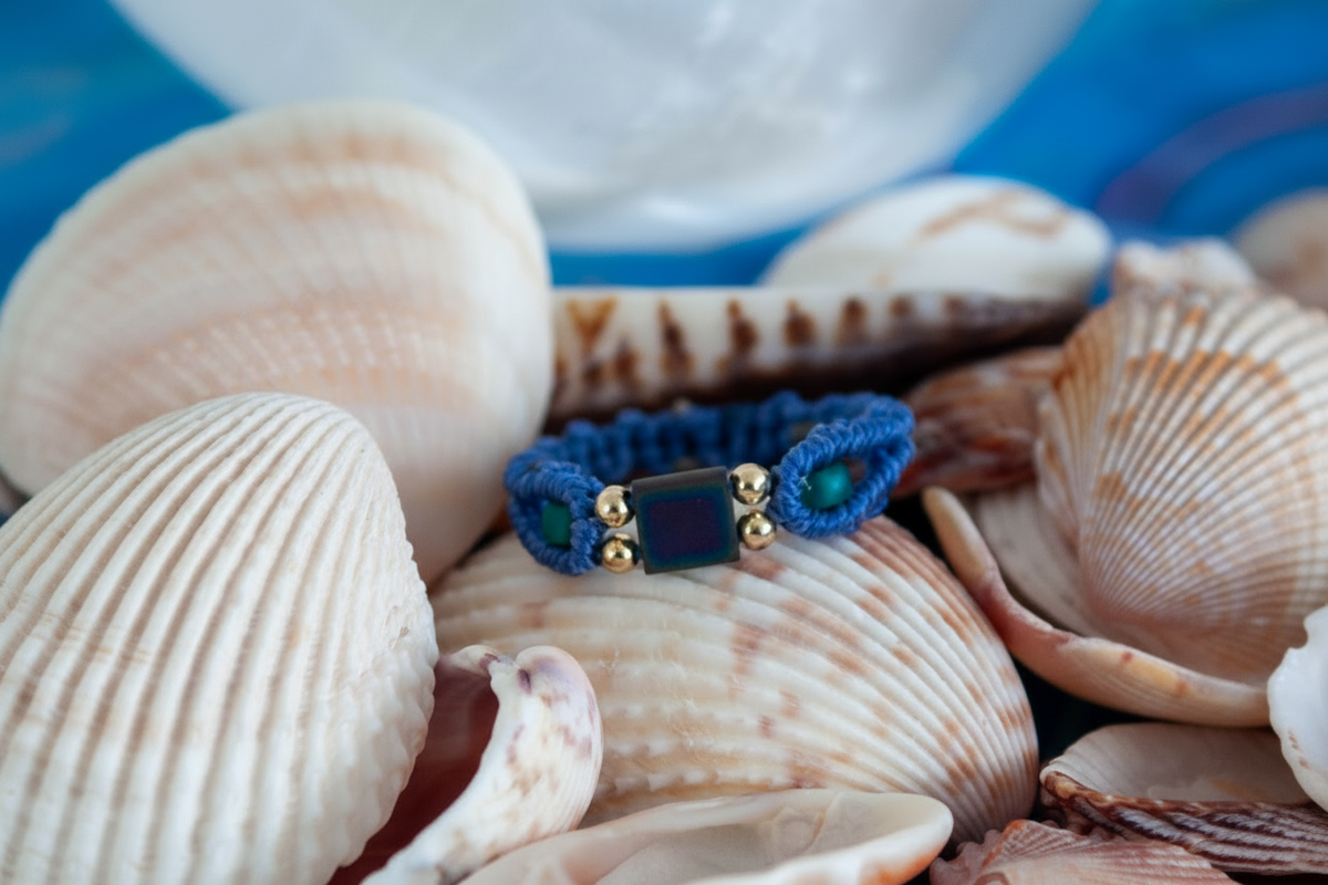 A close-up of a handmade minimalist periwinkle macrame ring with a square Miyuki Tila bead center, teal seed beads, and gold brass accent beads, nestled among white seashells with a selenite sphere in the background.