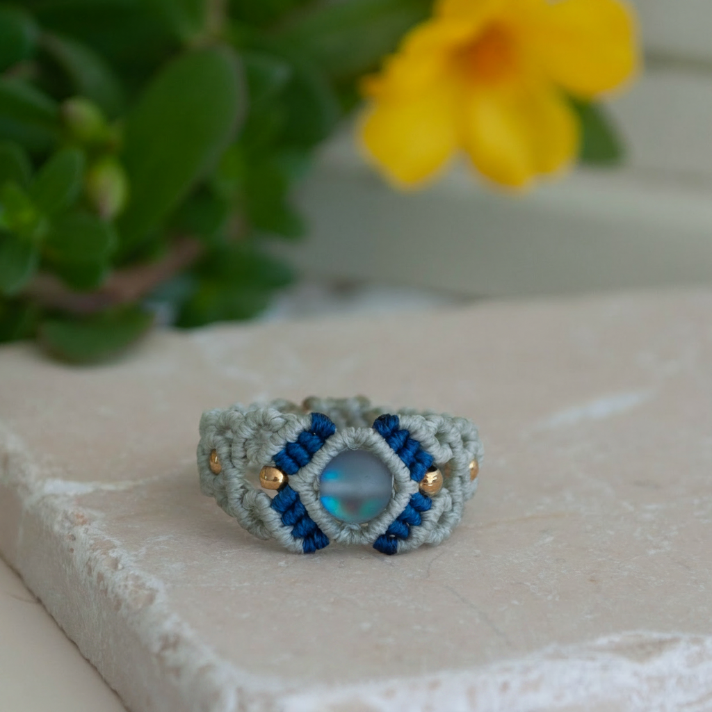 A handmade two-tone light gray and navy blue macrame cigar band ring with a shimmering iridescent mermaid glass bead at the center and gold brass accent beads, displayed on a white stone tile with a yellow flower and greenery in the soft-focus background