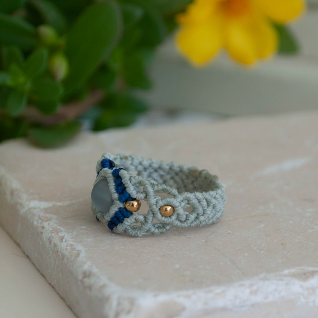 The side and back view of a handmade two-tone light gray and navy blue macrame cigar band ring showing the intricate knotwork band construction and gold brass accent beads, displayed on a white stone tile with a yellow flower in the soft-focus background