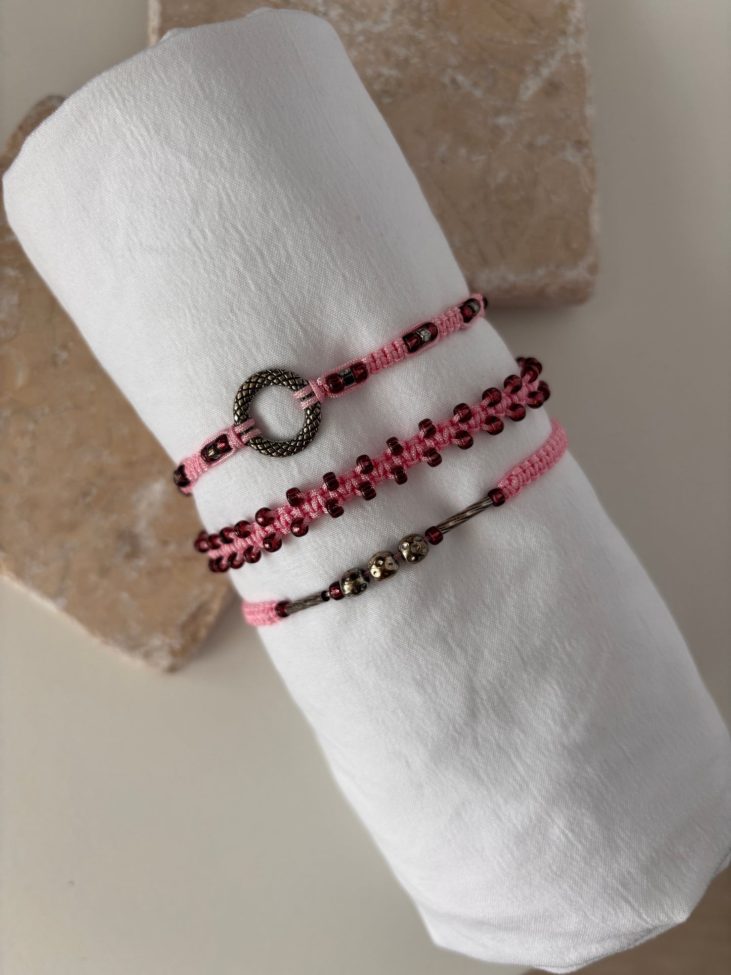 Three pink macrame bracelets displayed on a white linen roll against a stone background — one with a silver-tone open ring centerpiece, one with cranberry seed beads woven along the edges, and one with a silver-tone tube bead and silver-tone ball accent beads on pink nylon cord.