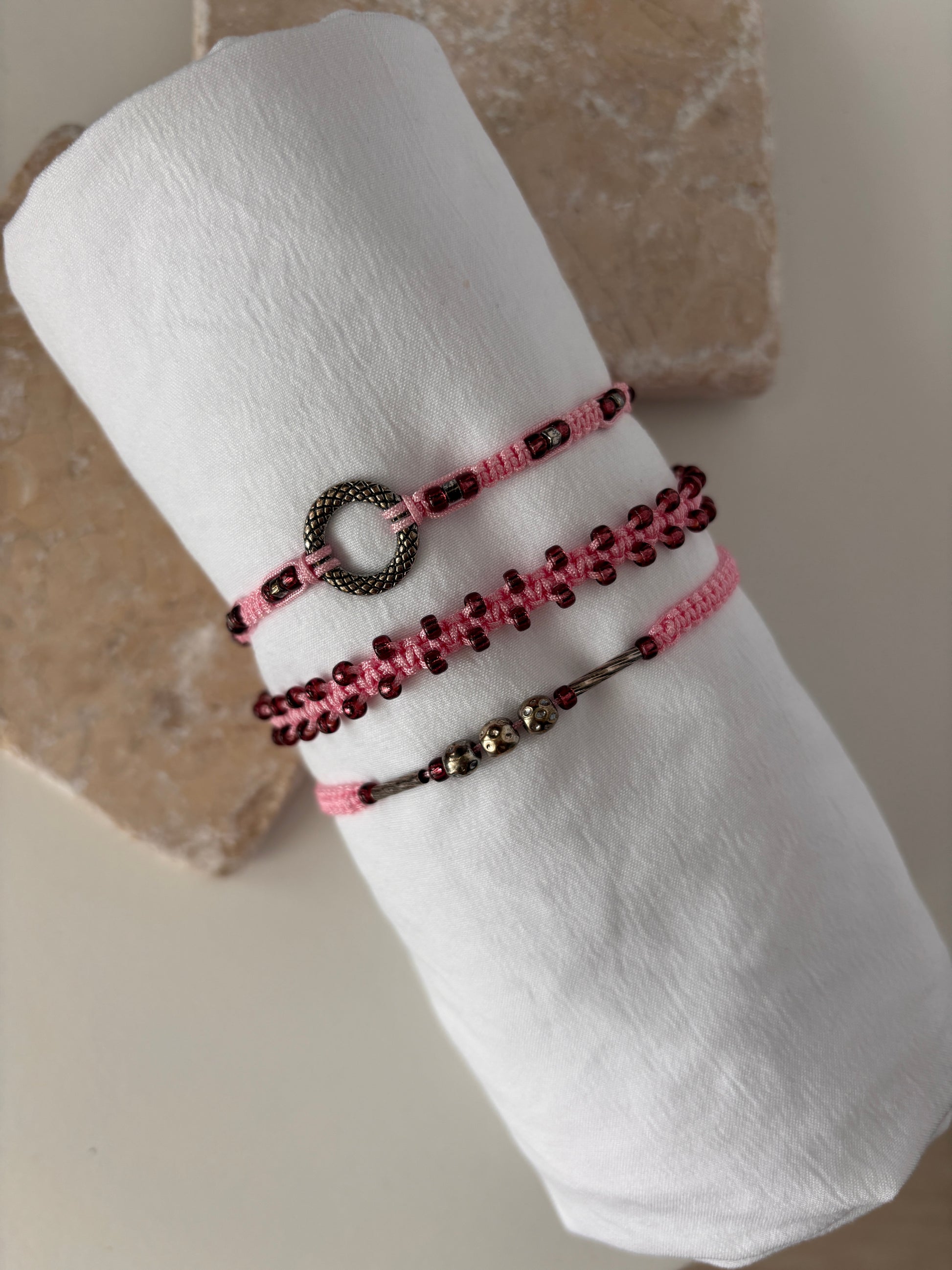 Three pink macrame bracelets displayed on a white linen roll against a stone background — one with a silver-tone open ring centerpiece, one with cranberry seed beads woven along the edges, and one with a silver-tone tube bead and silver-tone ball accent beads on pink nylon cord.