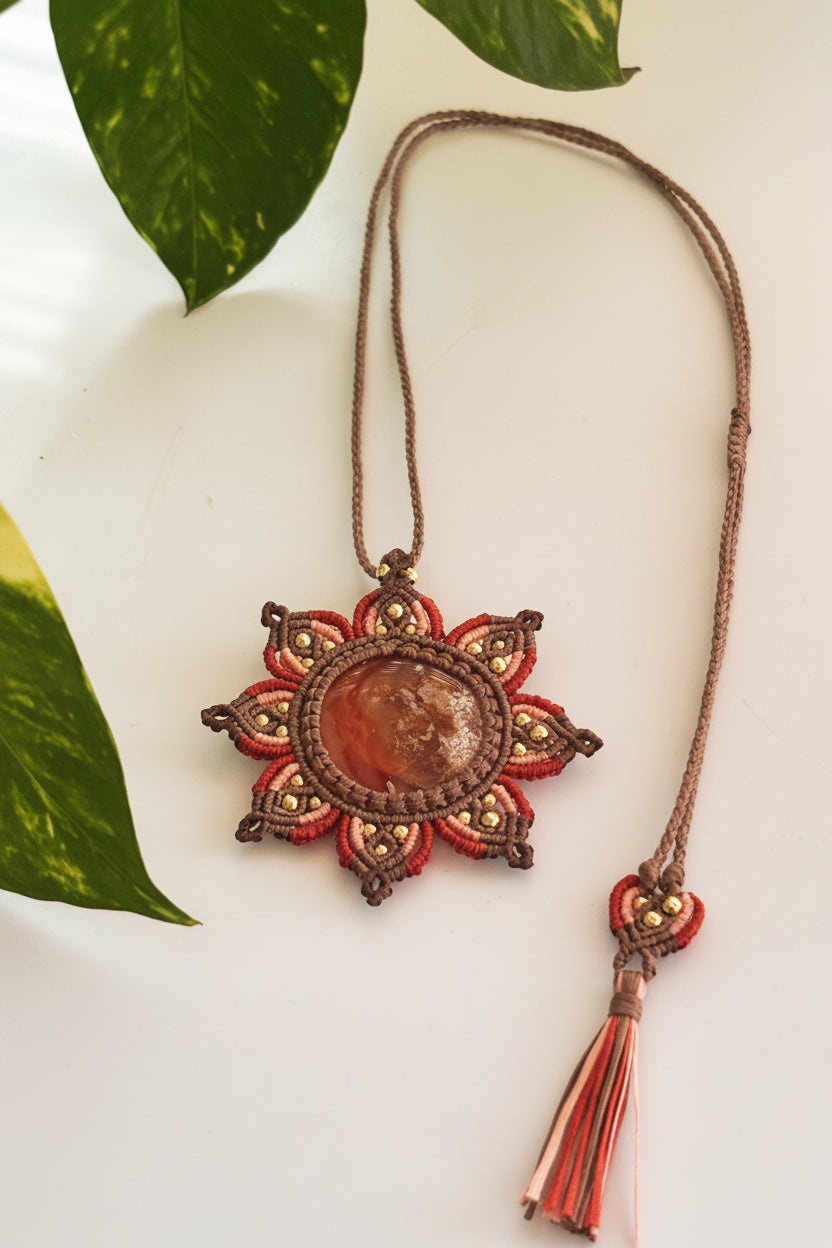 A handmade macrame pendant necklace featuring a central smooth Carnelian cabochon (amber/brown stone) surrounded by rust, peach, and brown woven cords with brass bead accents, displayed on a white surface with green leaves in the corner.