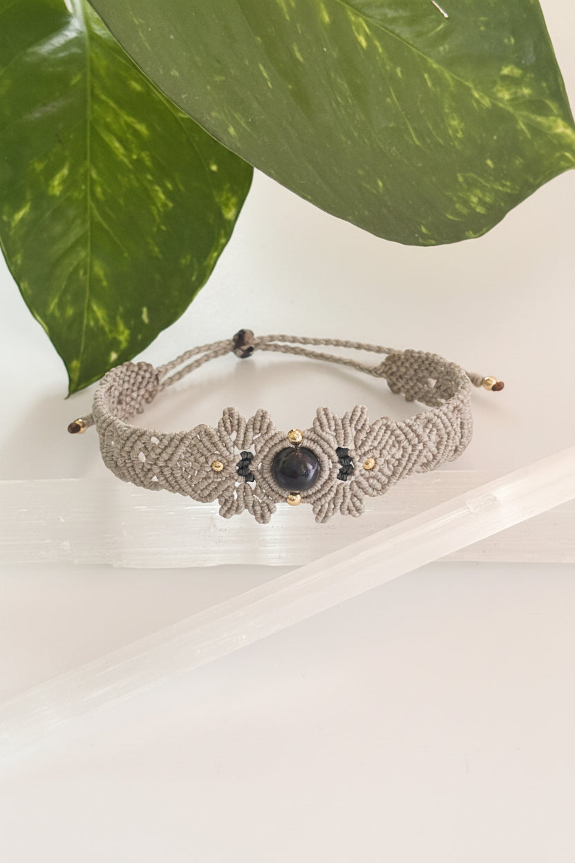 A handmade gray macrame bracelet with a central lapis lazuli stone and small brass beads, displayed on a clear acrylic block with green leaves in the background.