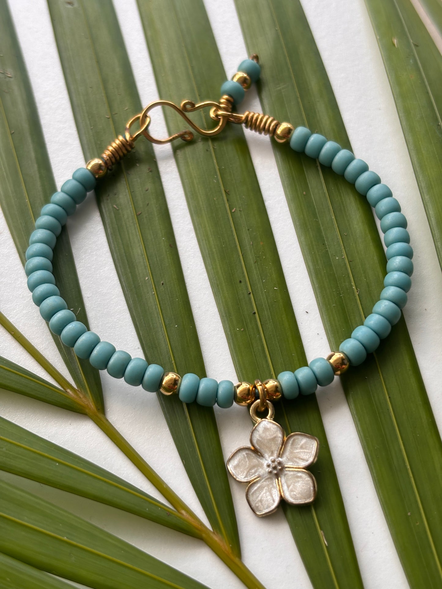 A beaded bracelet with turquoise blue seed beads, brass spacer beads, a handcrafted brass clasp, and a white enamel flower charm, displayed on green palm leaves over a white background.