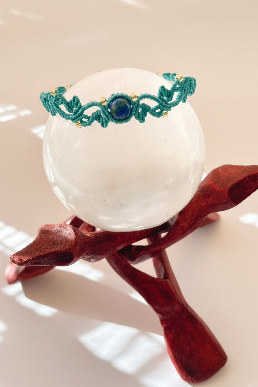 A handmade teal macrame bracelet with a central blue gemstone and gold-lined seed beads, displayed on a white crystal sphere atop a dark wooden stand.