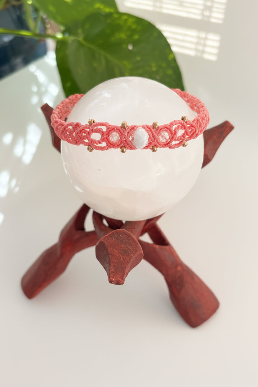 A salmon pink macrame bracelet with small brass beads and a faceted iridescent central crystal framed by small iridescent pink round faceted beads, displayed on a white crystal sphere atop a dark reddish-brown wooden stand.