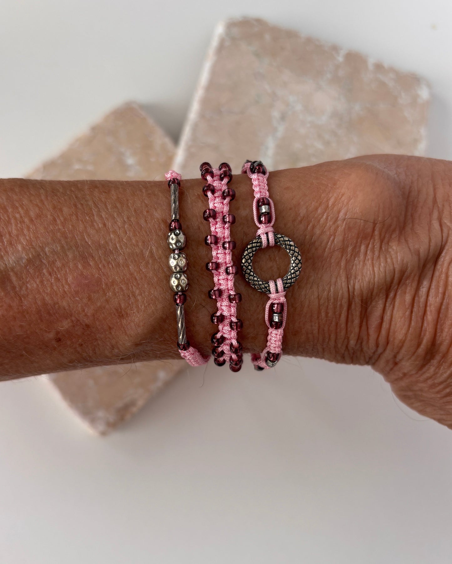 Three pink macrame bracelets stacked on a wrist against a white stone background — one with a silver-tone open ring centerpiece, one with cranberry seed beads woven along the edges, and one with a silver-tone tube bead and silver-tone ball accent beads on pink nylon cord