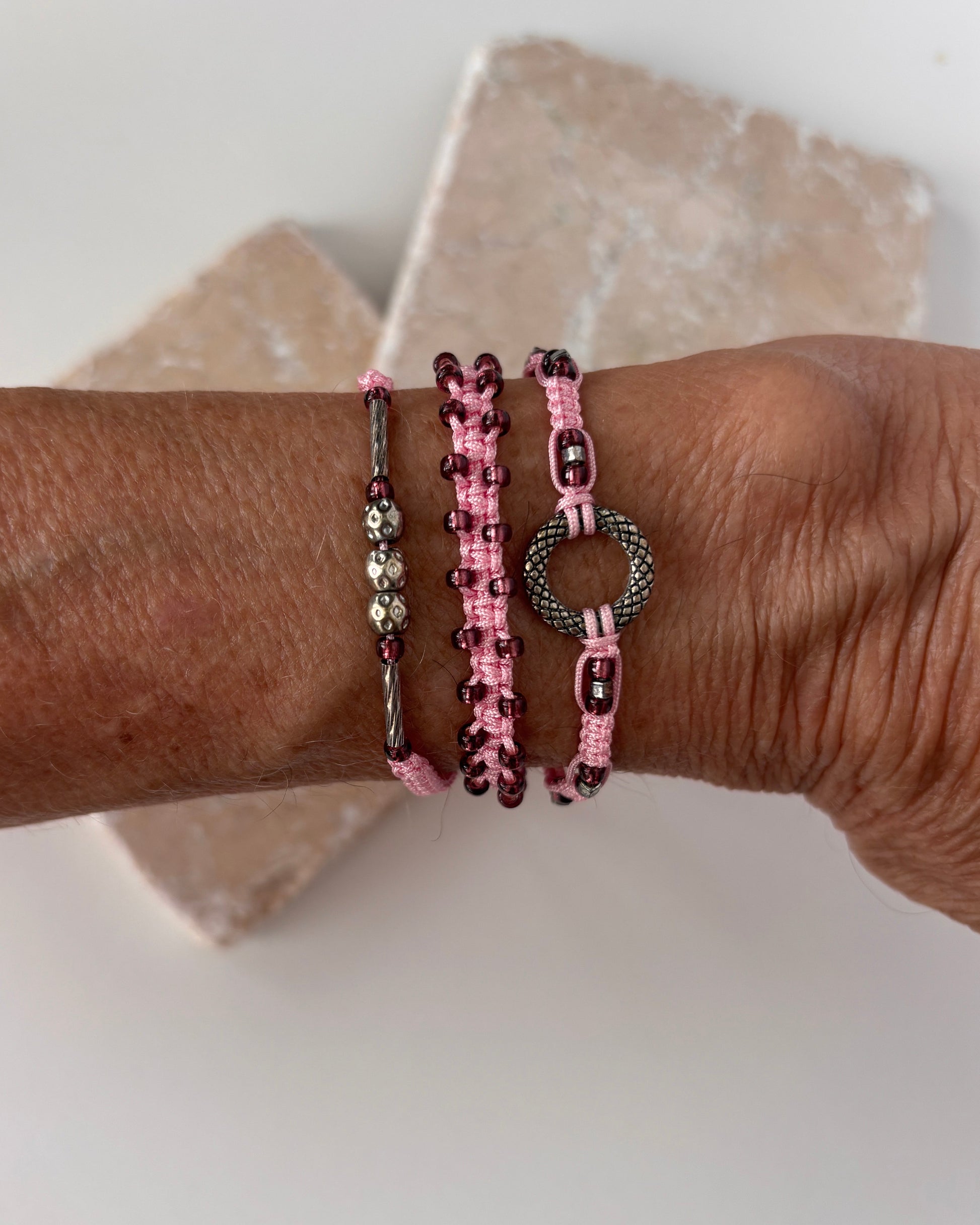 Three pink macrame bracelets stacked on a wrist against a white stone background — one with a silver-tone open ring centerpiece, one with cranberry seed beads woven along the edges, and one with a silver-tone tube bead and silver-tone ball accent beads on pink nylon cord