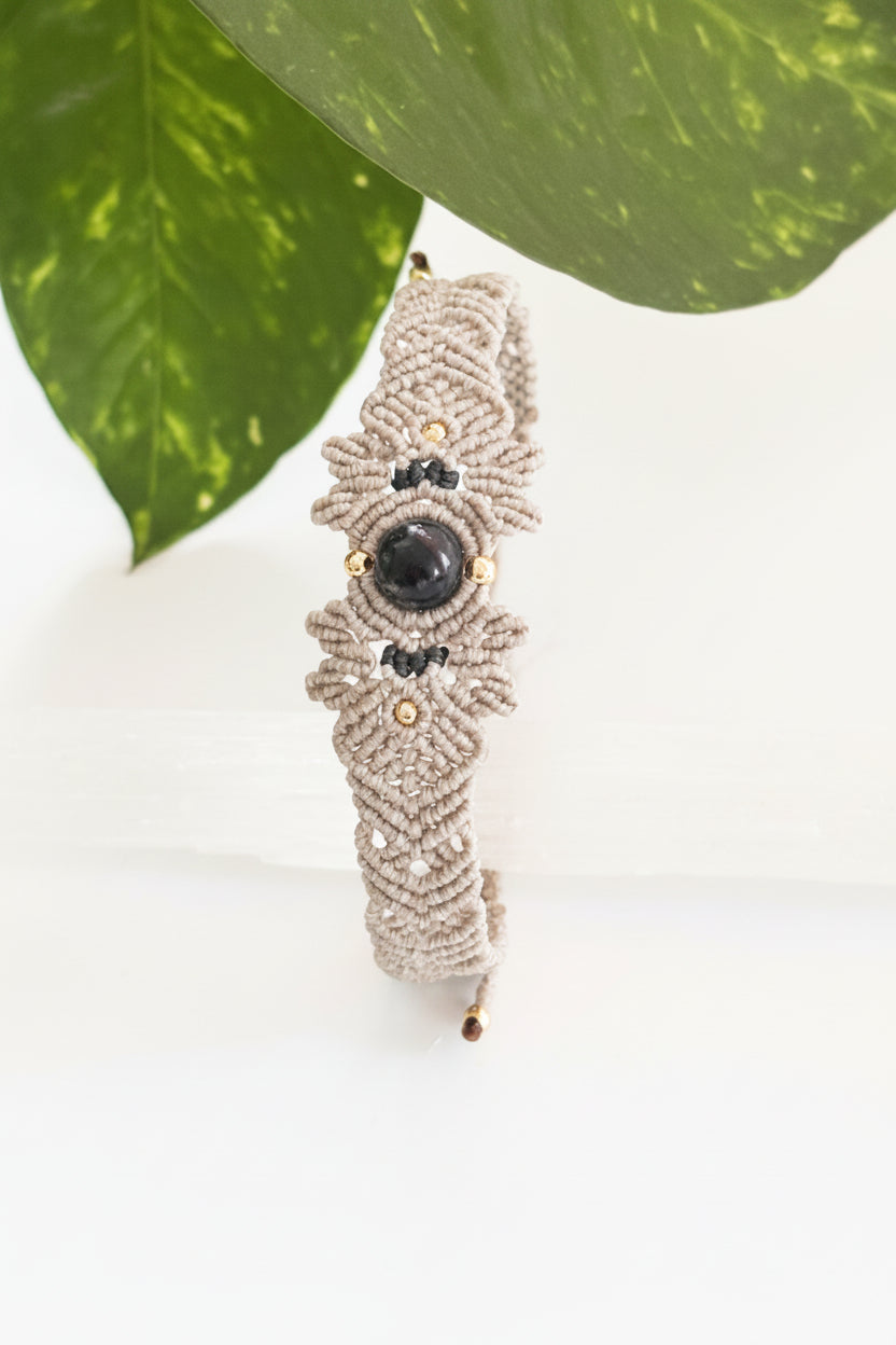 A handmade macrame bracelet in light gray with a central lapis lazuli stone and small brass accent beads, displayed vertically on a clear acrylic block with green leaves in the background.