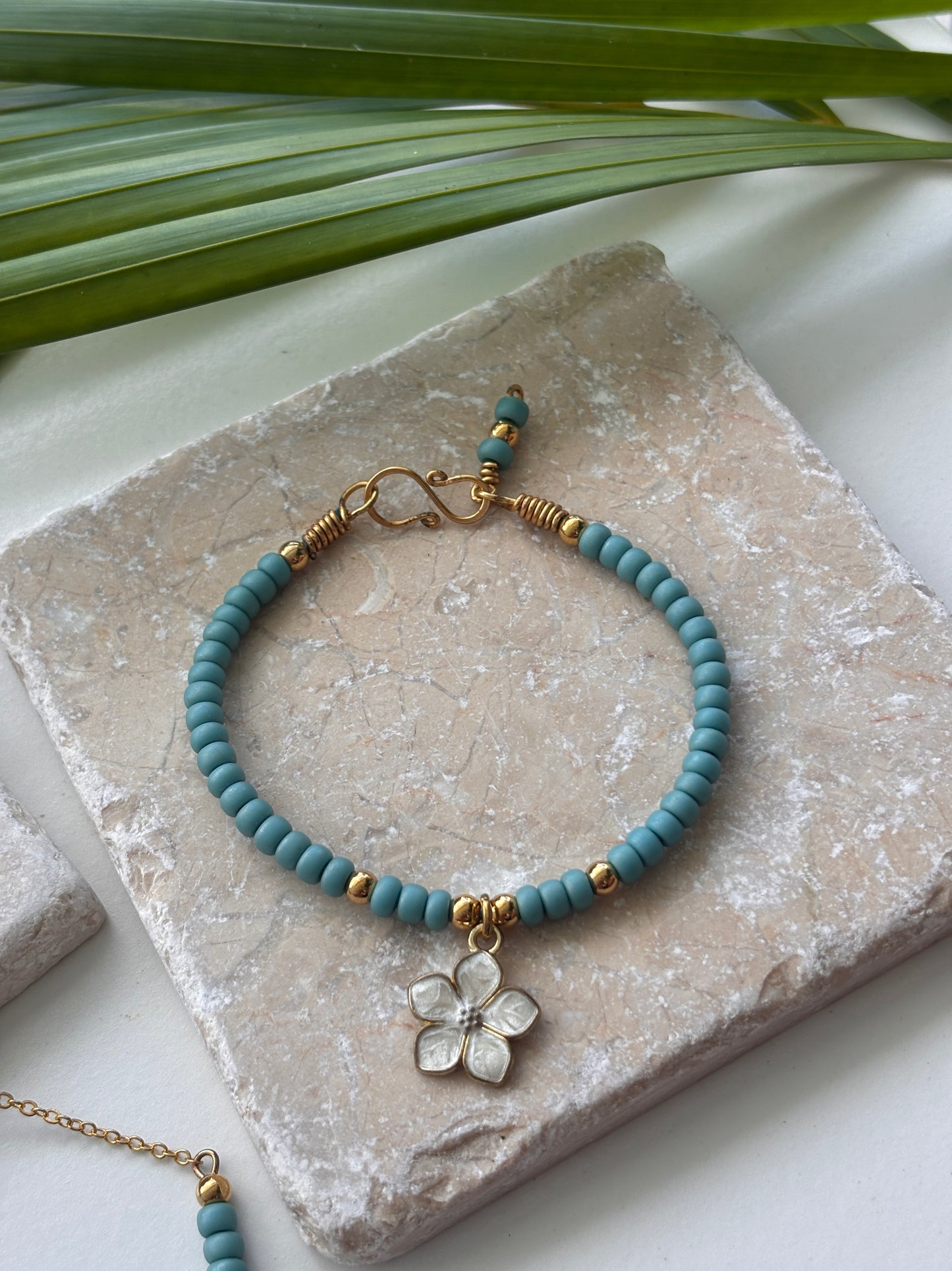 A turquoise blue beaded bracelet with brass hardware and a white enamel flower charm, displayed on a textured stone slab with green leaves in the background.