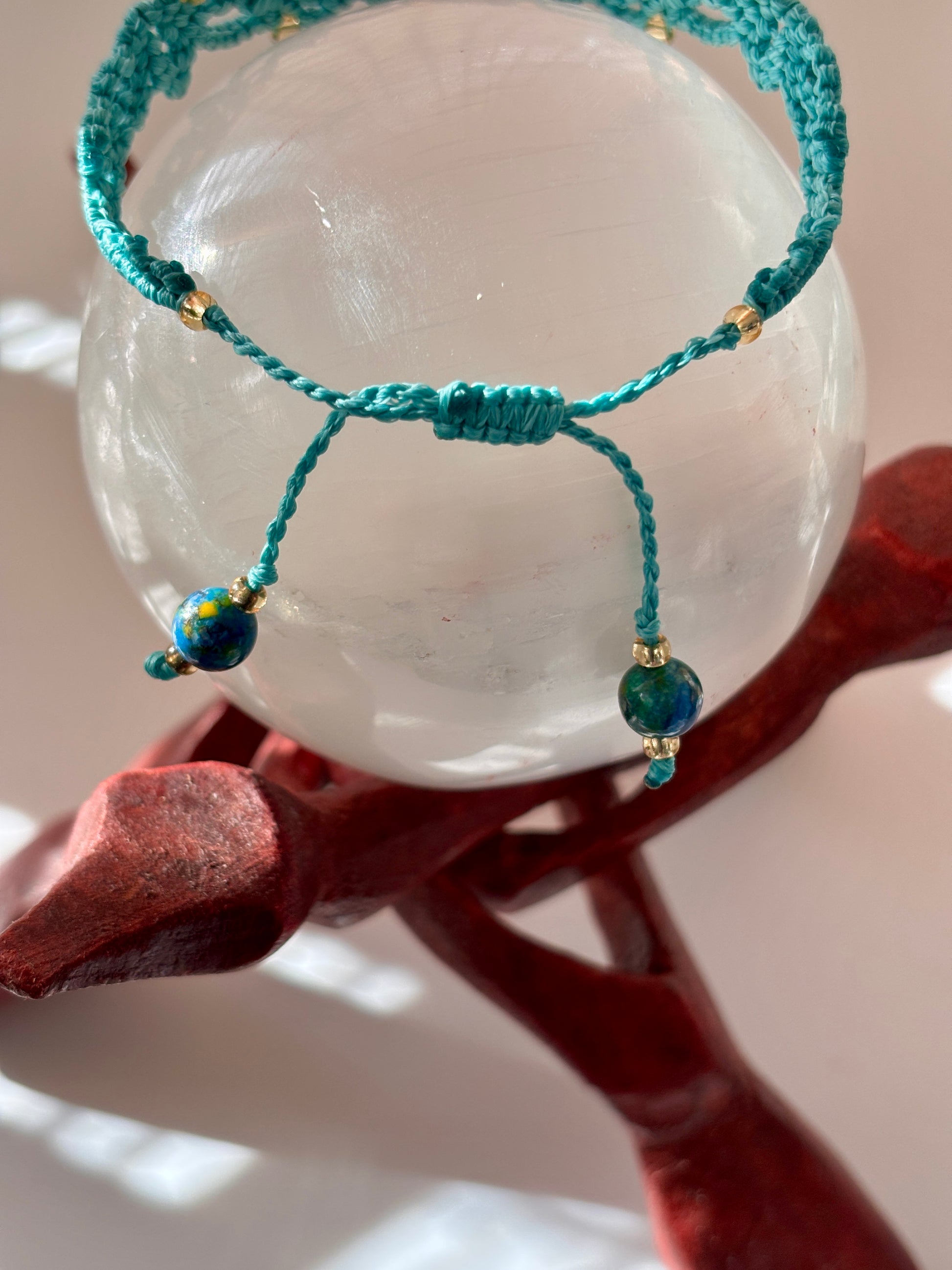 A teal macrame bracelet with small gold-lined seed beads and two dangling blue marbled beads, displayed on a clear glass sphere over a dark red wooden stand.