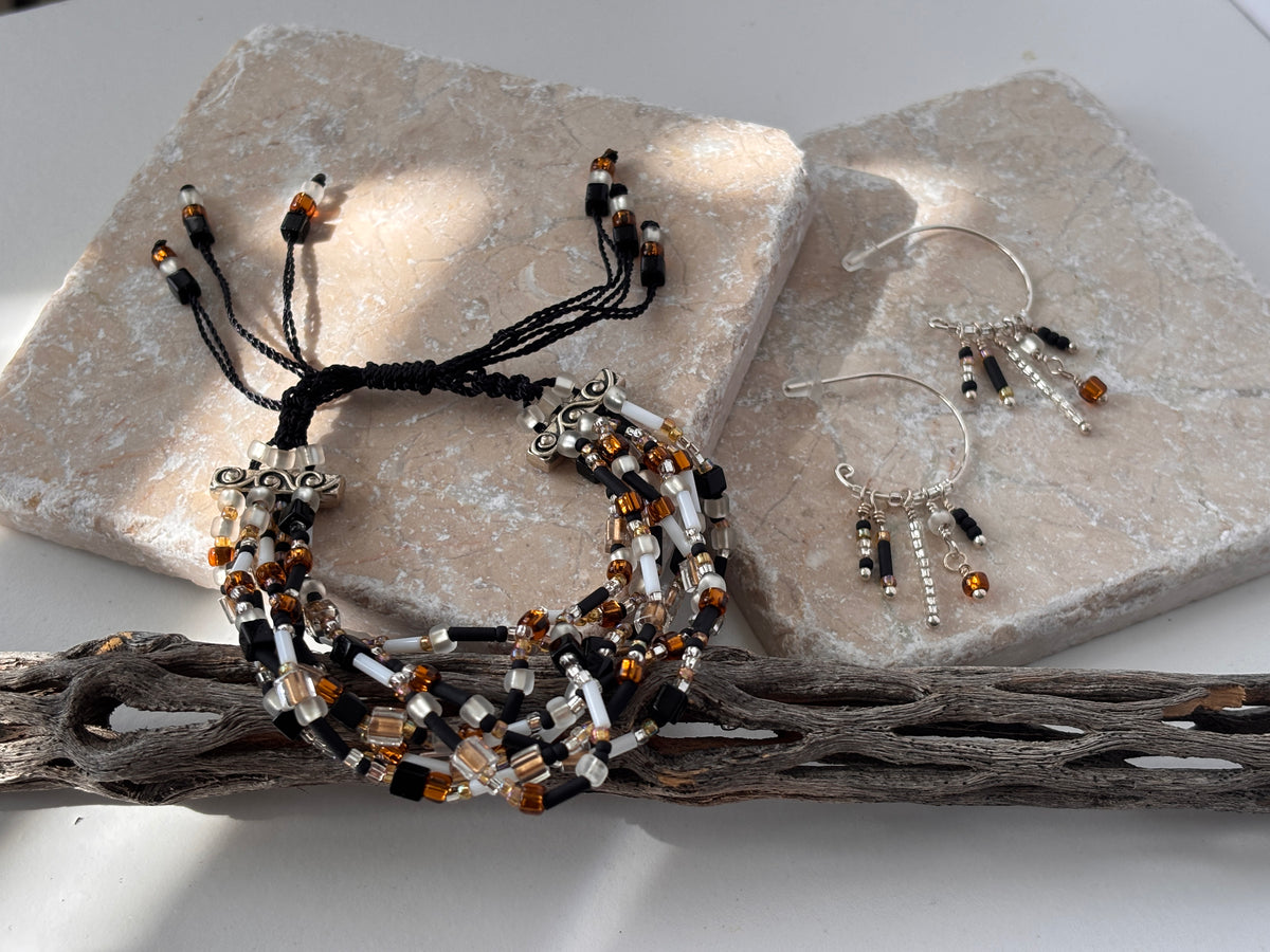 A handmade seven strand seed bead bracelet in black nylon macrame cord with mixed amber, white, and clear glass seed beads, cube beads, and bugle beads with silver-tone scroll spacer bars, styled with matching silver-tone hoop earrings with beaded dangles, displayed on overlapping pink stone slabs with driftwood on a white surface.  Items sold separately.