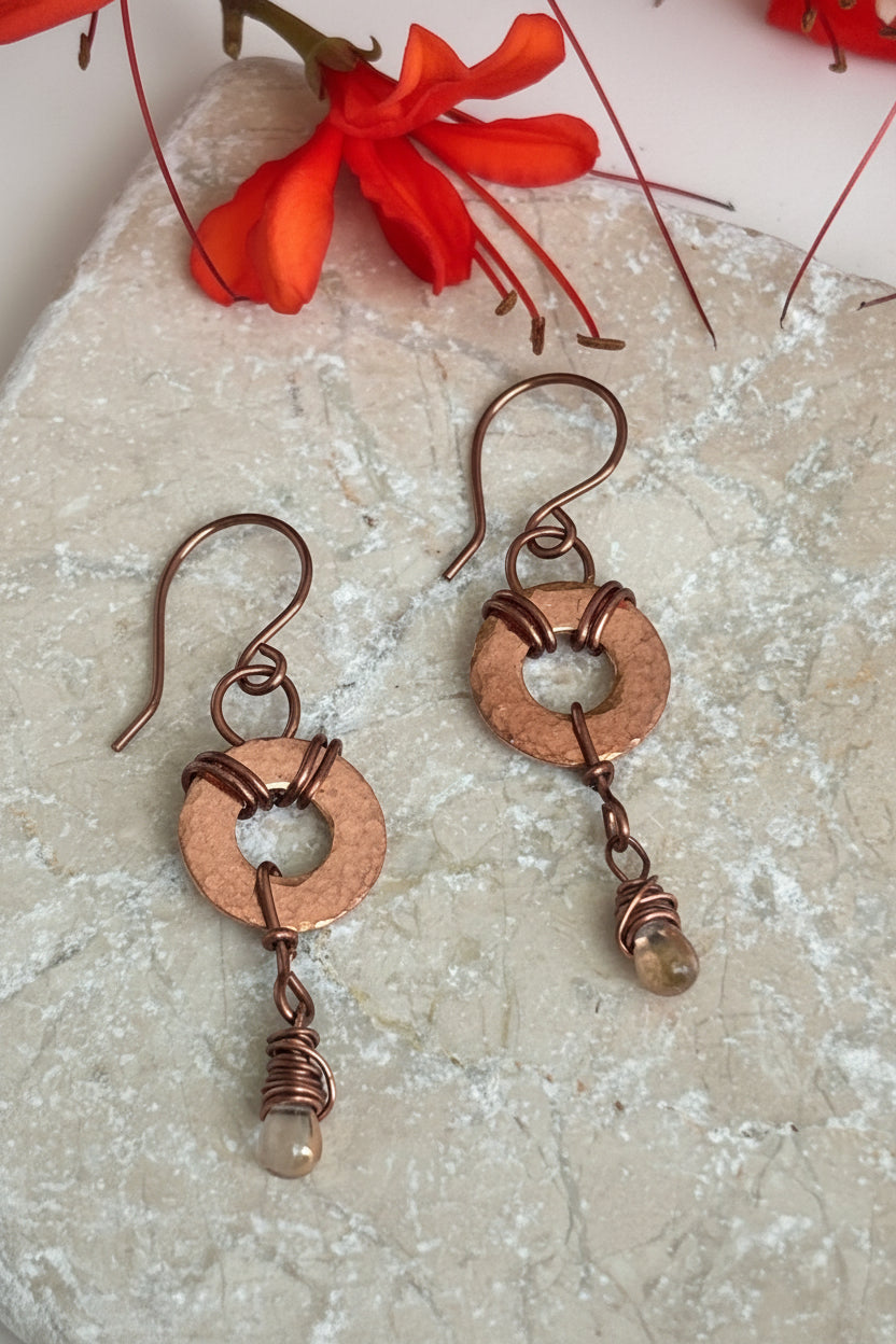 A pair of handmade copper drop earrings with waterdrop wire-wrapped beads, displayed on a textured light stone surface with red flower stems in the background.