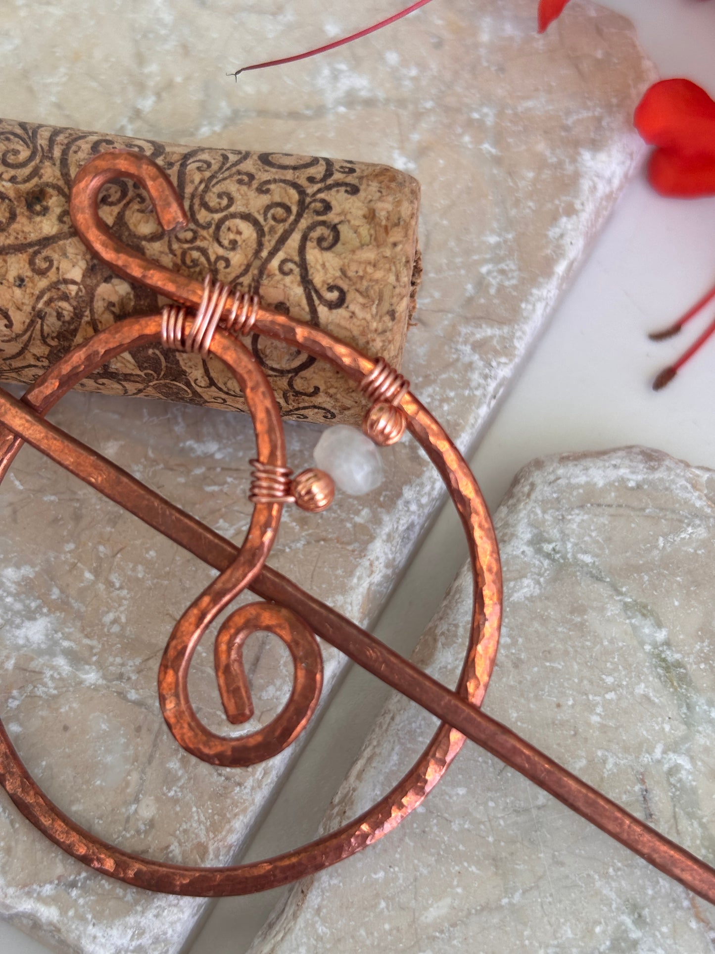 A hammered copper hair stick with wrapped wire details with copper and moonstone beads, shown resting on a cork and textured stone surface.