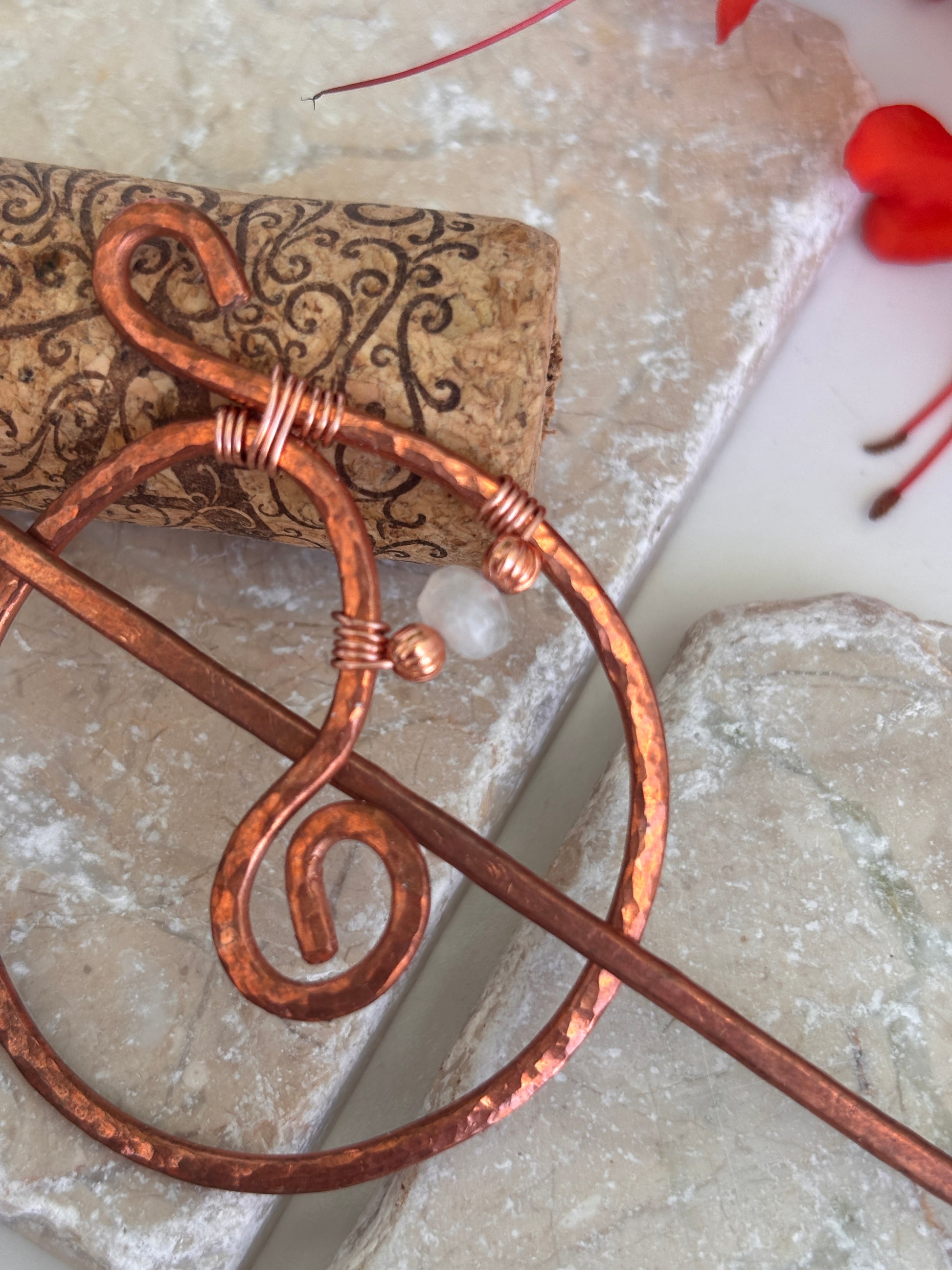 A hammered copper hair stick with wrapped wire details with copper and moonstone beads, shown resting on a cork and textured stone surface.