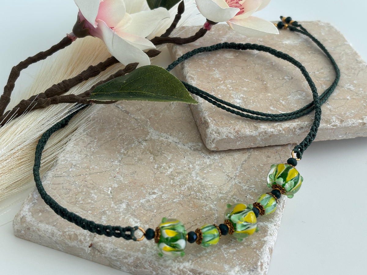 Clear yellow, green, and white swirl lampwork necklace with dark green macrame cord, styled on travertine tile.