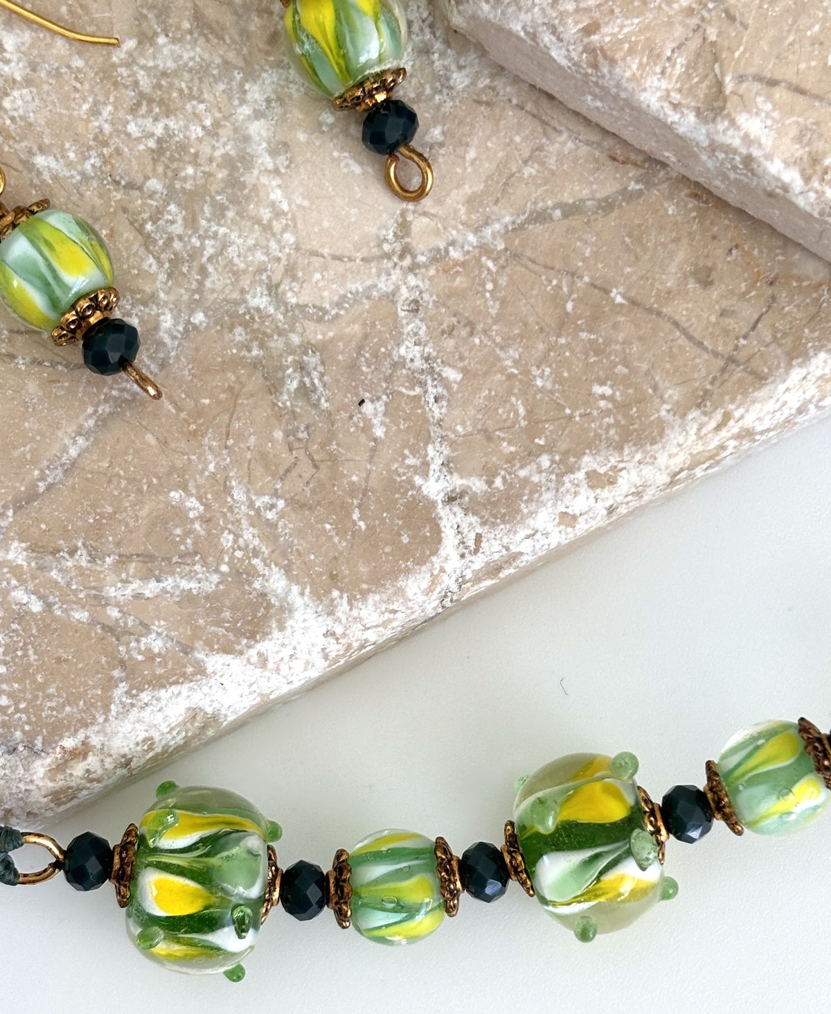 Close-up detail of handmade dark green macrame necklace showing 10×14mm textured tropical swirl lampwork glass beads with raised glass dot detail, gold-tone daisy spacer beads, and 3mm dark green faceted crystal rondelles, with coordinating lampwork glass drop earrings on travertine tile.