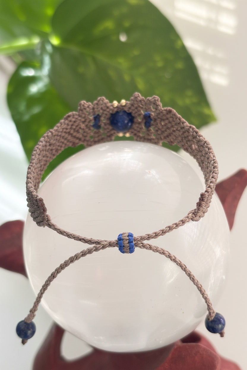 The backside of a handmade macrame bracelet in a brown cord, decorated with brass beads and a central Lapis Lazuli stone and a macrame bead slide closure displayed on a clear glass sphere.