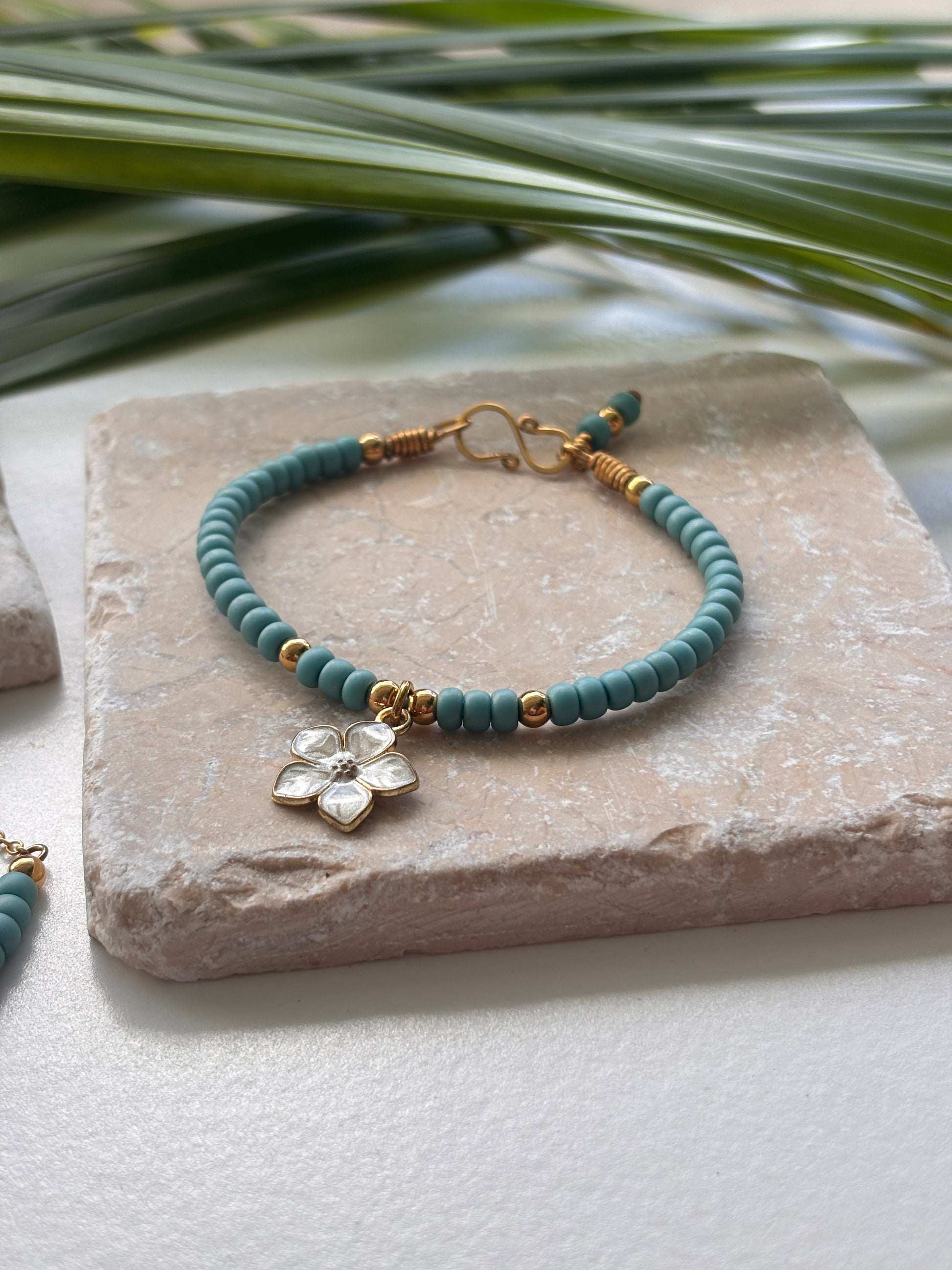 A turquoise blue beaded bracelet with brass hardware and a small white enamel flower charm, displayed on a textured stone slab with green leaves in the background.