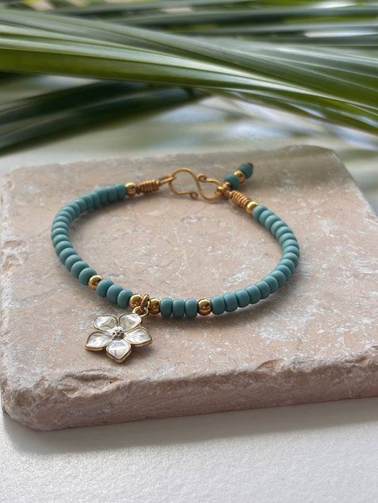 Turquoise 6/0 matte seed bead bracelet with 4mm brass beads and an ivory enamel flower charm displayed on a textured stone slab with green leaves.